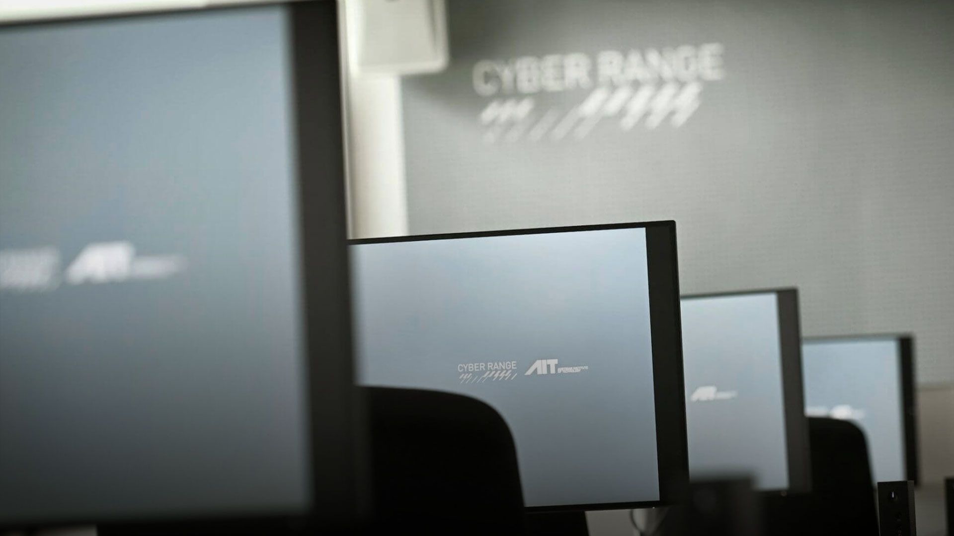 Mehrere Computerbildschirme in einem Raum mit der Aufschrift 'CYBER RANGE' an der Wand im Hintergrund