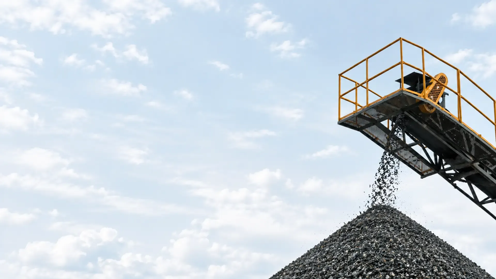 Mining conveyor depositing gravel onto a large stockpile beneath a pale blue sky.