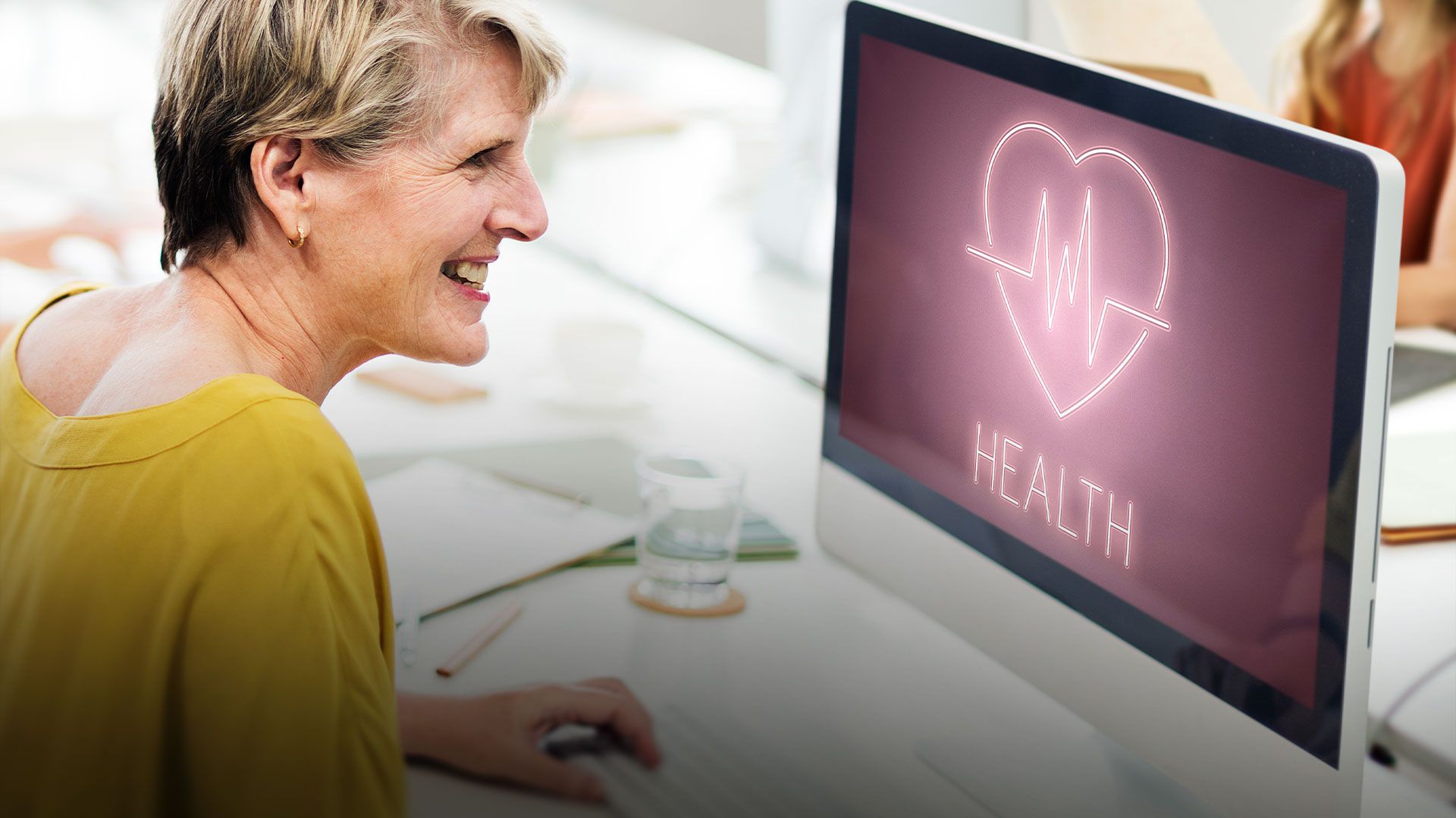 Ältere Frau lächelt am Schreibtisch vor Computerbildschirm mit Herz und Herzfrequenzsymbol und dem Wort 'HEALTH'