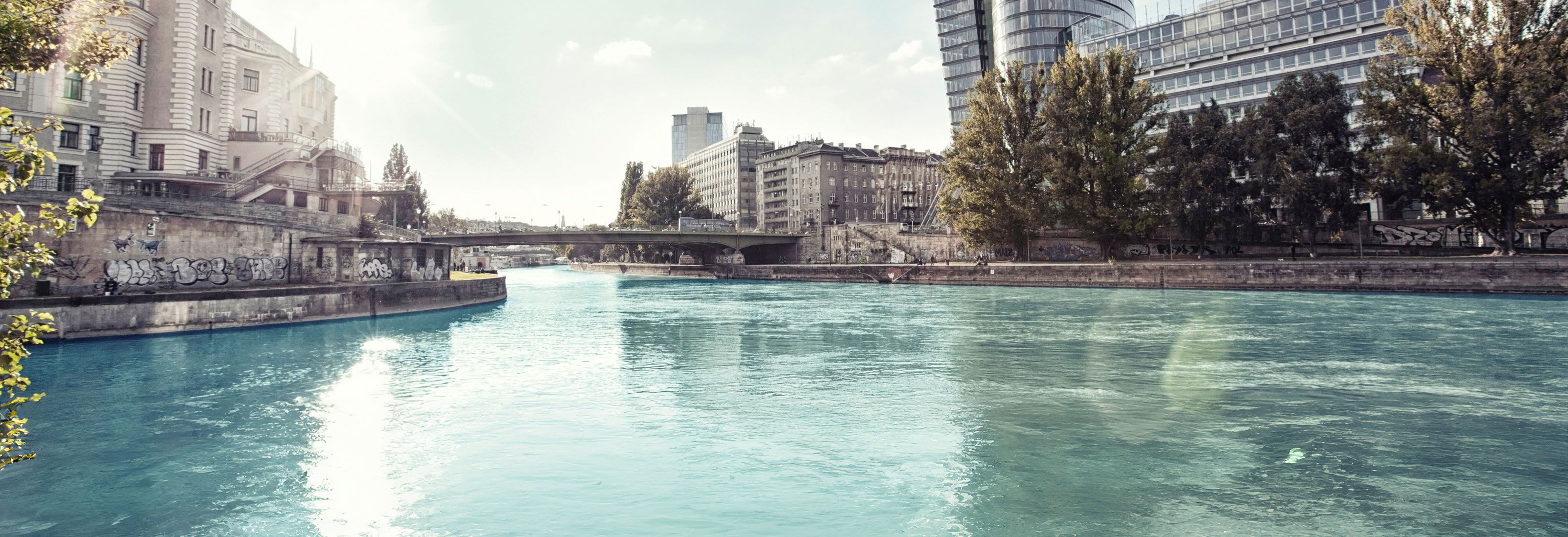 Fluss mit klarem Wasser, umgeben von Bäumen und modernen sowie historischen Gebäuden unter blauem Himmel mit Wolken.