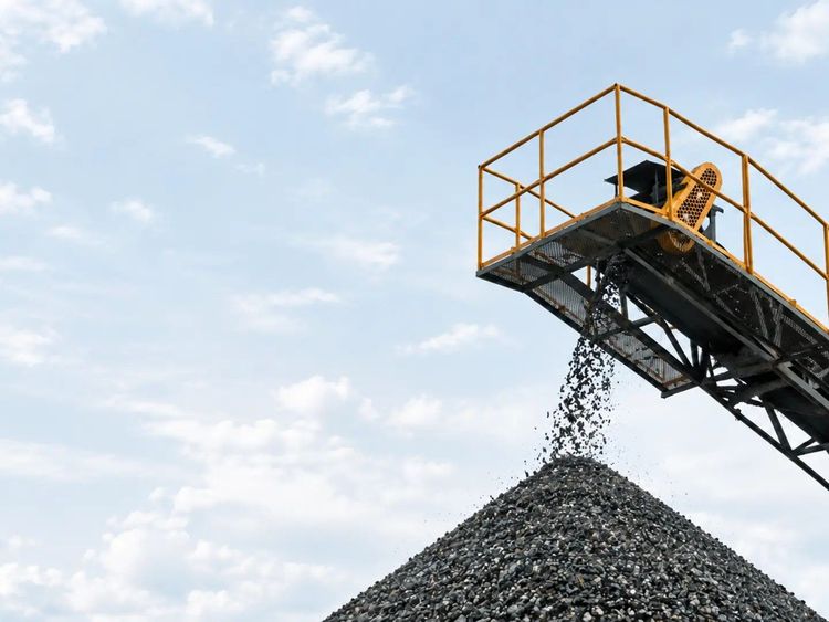 Mining conveyor depositing gravel onto a large stockpile beneath a pale blue sky.