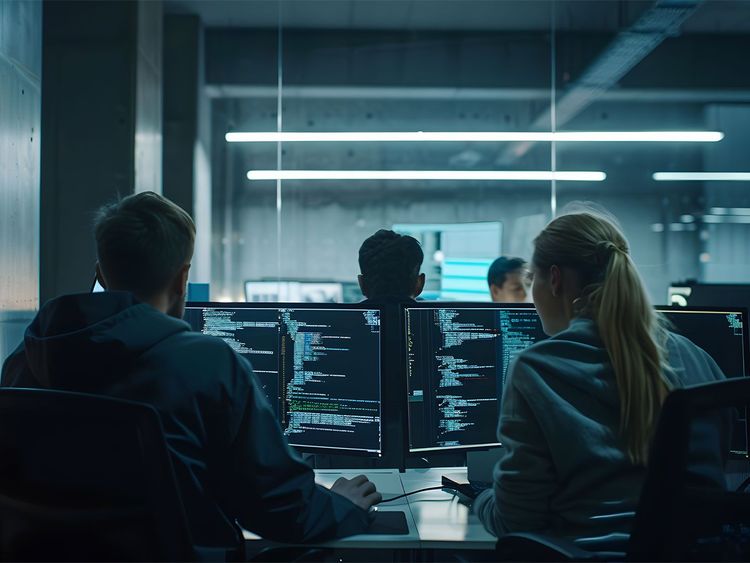 Drei Personen sitzen vor mehreren Monitoren mit Programmiercode in einem dunklen Büro.