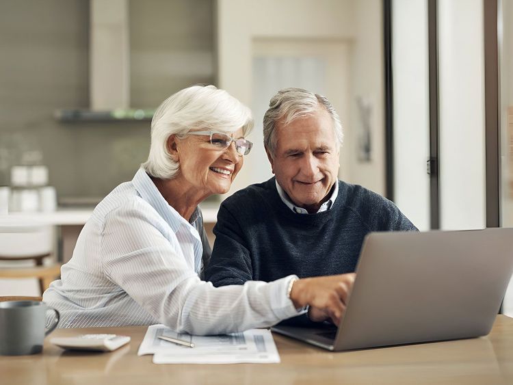 Älteres Paar sitzt am Tisch und schaut lächelnd auf einen Laptop, Frau zeigt auf den Bildschirm